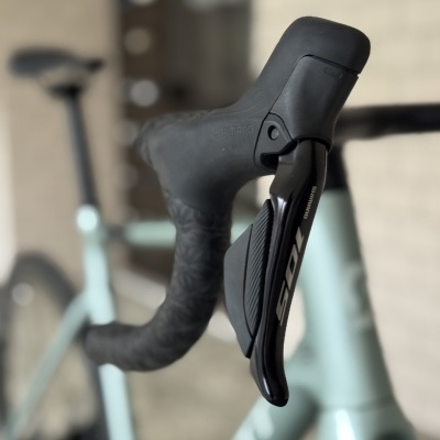 Punho de guiador de bicicleta preto com manete Shimano e bicicleta azul claro desfocada ao fundo.