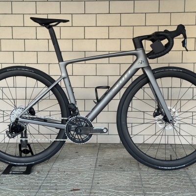Bicicleta de estrada cinza com rodas pretas apoiada num suporte diante de uma parede clara.
