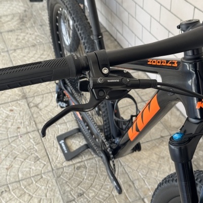 Bicicleta de montanha preta com detalhes laranja e manete Shimano no guiador.