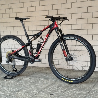 Bicicleta de montanha preta e vermelha com suspensão dianteira e pneus Maxxis encostada em parede de azulejos.