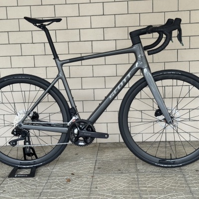 Bicicleta de estrada cinzenta SCOTT com pneus pretos e componentes shimano 105 encostada a parede de azulejos brancos