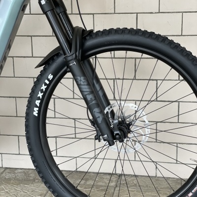 Roda dianteira de bicicleta com pneu Maxxis e suspensão preta contra parede de azulejos brancos