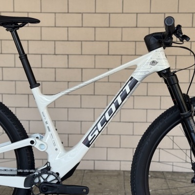 Bicicleta de montanha branca Scott com quadro carbono e suspensão preta encostada a parede de azulejos beje.