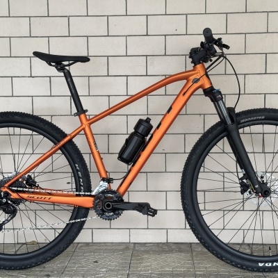 Bicicleta de montanha laranja com pneus pretos e garrafa de água, encostada em parede de tijolos brancos