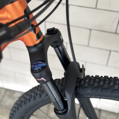 Bicicleta de montanha com suspensão dianteira preta e laranja, pneus robustos, em fundo de azulejos brancos e pavimento de lajes cinzentas.