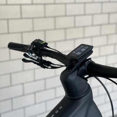 Bicicleta elétrica preta com ecrã digital no guiador e nome PATRON no quadro em fundo de azulejos brancos