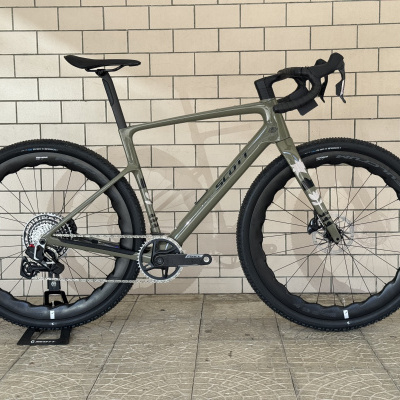 Bicicleta de corrida gravel Scott com quadro cinza e detalhes pretos encostada a parede de azulejos.