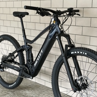 Bicicleta de montanha elétrica preta SCOTT com pneus MAXXIS junto a uma parede de azulejos brancos