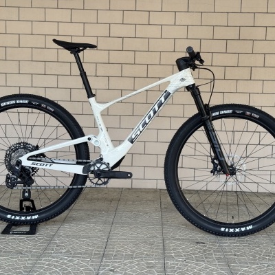 Bicicleta de montanha Scott branca e preta apoiada em suporte com parede de tijolos ao fundo