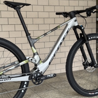 Bicicleta de montanha SCOTT cinza com pneus pretos e suspensão, em frente a uma parede de tijolos