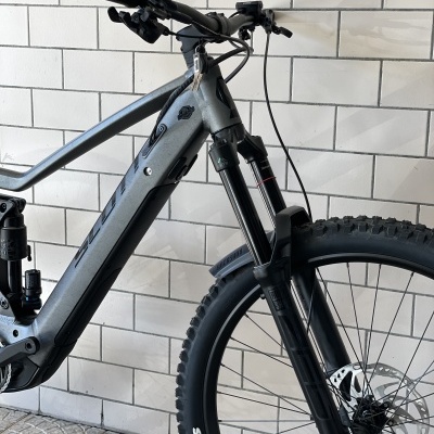 Bicicleta de montanha cinza e preto com pneus Maxxis junto a parede de azulejos brancos