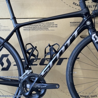 Bicicleta de estrada SCOTT preta com componentes Shimano 105 e roda Schwalbe em caixa de cartão.
