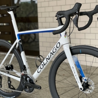 Bicicleta de estrada branca e azul Colnago com rodas pretas apoiada frente a parede de tijolos