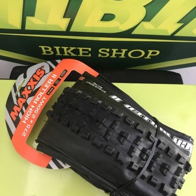 Pneu Maxxis High Roller II preto com etiqueta laranja e branca sobre superfície verde, cartaz BIKE SHOP ao fundo