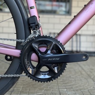 Pedaleira preta Shimano 105 na bicicleta rosa metálica com corrente cinza e fundo de azulejos