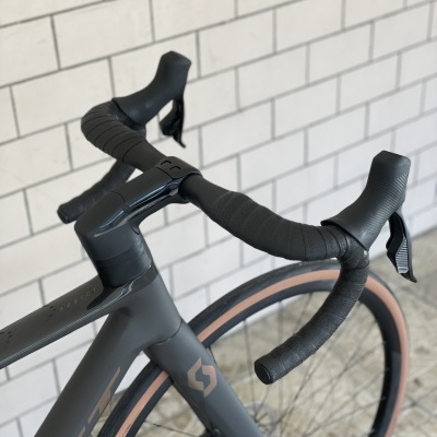 Bicicleta de estrada cinza escuro com guiador preto, pneus com borda castanha e parede de tijolos ao fundo