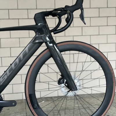 Bicicleta de estrada preta com detalhes cinza e pneus castanhos junto a parede de azulejos brancos.