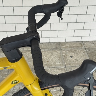 Guiador preto com fita preta e quadro amarelo com autocolante Danibikes, manetes Shimano, fundo de azulejos brancos.
