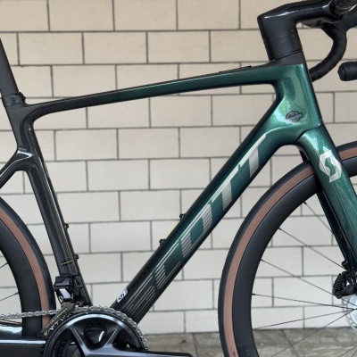 Bicicleta de estrada Scott verde metálico com componentes pretos e logótipo branco junto a parede de azulejos.