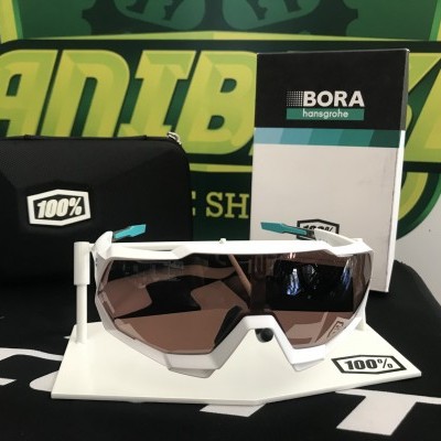 Óculos de desporto 100% com armação branca e lentes fumadas sobre base branca, saco preto e embalagem BORA hansgrohe ao fundo