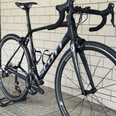 Bicicleta de estrada preta SCOTT com pneus SCHWALBE e sistema Shimano Ultegra encostada a uma parede de azulejos.