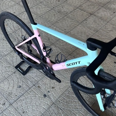 Bicicleta de estrada Scott em azul claro e rosa com selim e guiador pretos numa calçada de pedra.