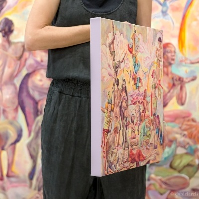 Pessoa segura tela colorida com pintura de figuras humanas e elementos surrealistas em frente a mural com figuras femininas