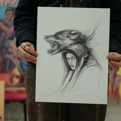 Desenho a carvão de mulher com capuz de pele de lobo, segura por pessoa em estúdio de arte com pinturas ao fundo