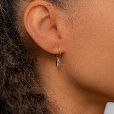 Brinco pequeno dourado com pedra transparente usado no lóbulo da orelha.