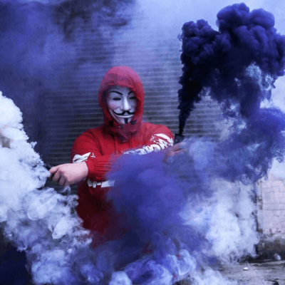Pessoa mascarada com casaco vermelho segurando flares de fumo branco e azul num ambiente urbano