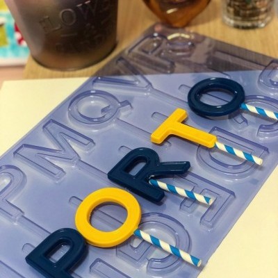 Tabuleiro azul com letras coloridas formando 'PORTO' e vaso com planta ao fundo