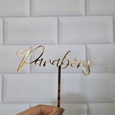Cake topper dourado com texto 'Parabéns' segurado à frente de uma parede branca de azulejos.