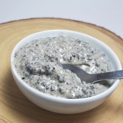 Recipiente branco com creme cinzento claro com sementes pretas e colher metálica sobre base de madeira