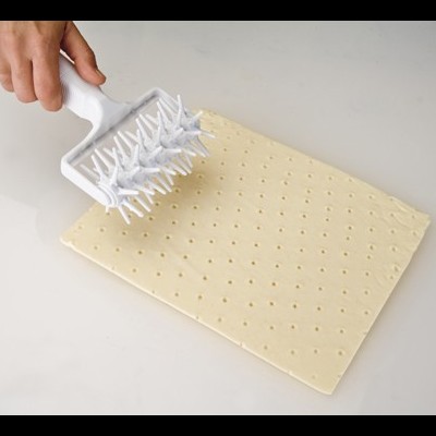Folha de massa retangular perfurada com rolo de pinos branco numa superfície clara.