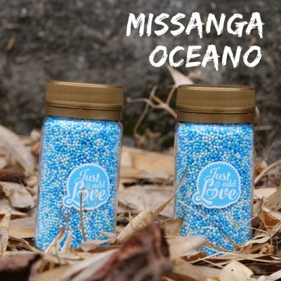 Dois frascos com missangas azuis e brancas, rótulo 'Just add Love', texto 'MISSANGA OCEANO' ao fundo