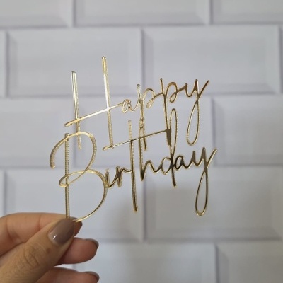 Decoração dourada de acrílico com 'Happy Birthday' segurada por mão feminina