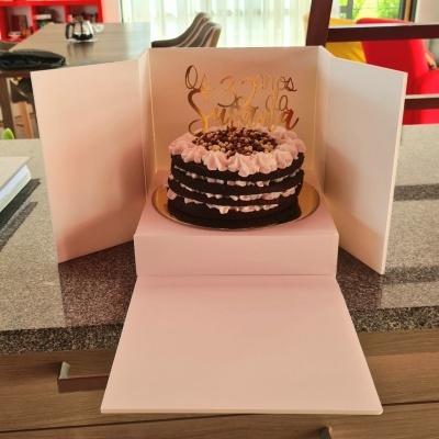 Bolo de chocolate com cobertura rosa e topper dourado dentro de caixa branca aberta