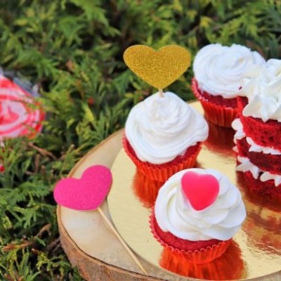 Cupcakes vermelhos com cobertura branca e corações decorativos em prato dourado sobre base de madeira com folhagem ao fundo.