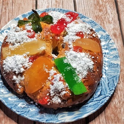 Bolo-rei decorado com frutas cristalizadas e açúcar em pó num prato azul sobre mesa de madeira