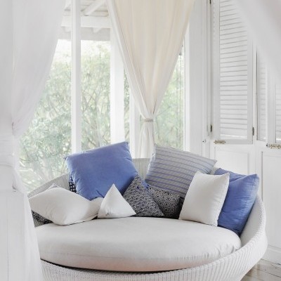 Sofá redondo em rattan branco com almofadas coloridas junto a janela com cortinas brancas