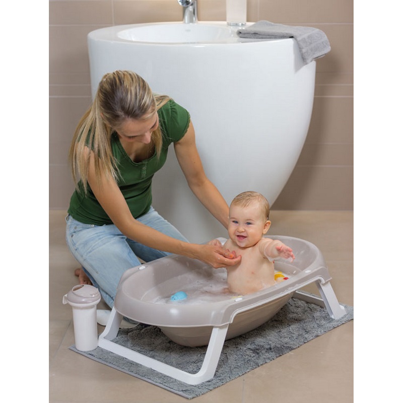 Banheira OKBaby Slim Bathtub Banheira de bebé cinzenta clara com pernas brancas sobre tapete cinzento, mulher a cuidar do bebé