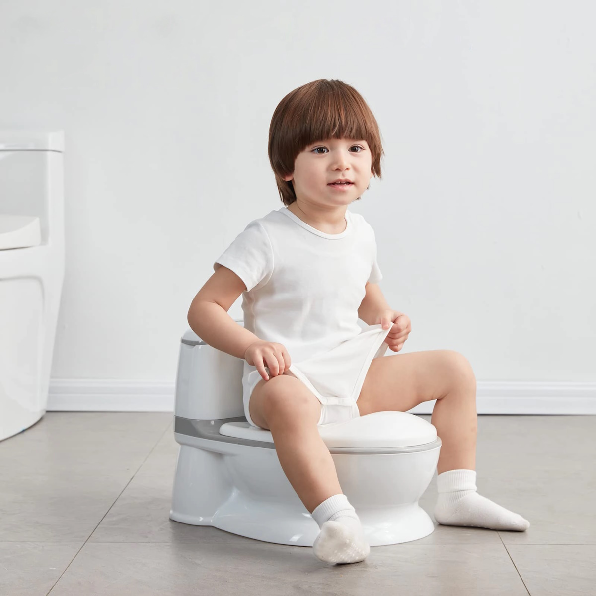 Sanita Kikkaboo Flushy Potty Toilet Seat Criança sentada num penico plástico branco com detalhe cinzento.