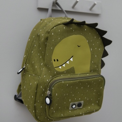 Mochila verde com imagem de dinossauro e bolso frontal pendurada num cabide branco