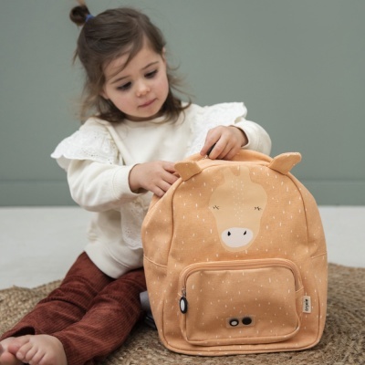 Mochila castanha clara com rosto de animal e chifres em tecido sobre tapete com criança ao fundo