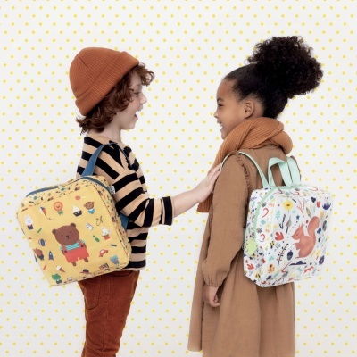 Duas crianças com mochilas coloridas e padrão infantil de animais, sorrindo em fundo de bolinhas amarelas.