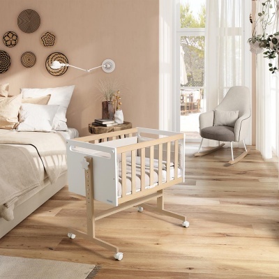 Berço de bebé em madeira clara e branca num quarto decorado com cama, almofadas, cestaria na parede e poltrona junto a janela