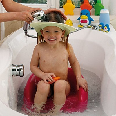 Criança num assento vermelho na banheira com chapéu de proteção para banho e brinquedos coloridos na borda.