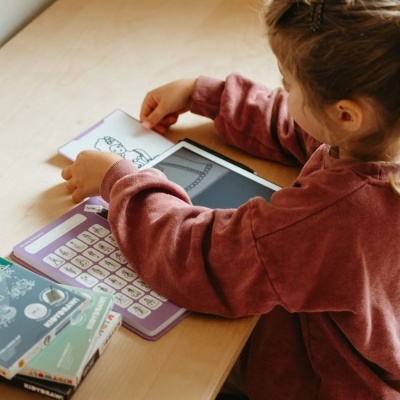 Criança com camisola castanha a usar tablet e livros com ilustrações numa mesa de madeira