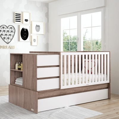 Berço de madeira clara com gavetas brancas e grades brancas, em quarto decorado com quadros e janela grande