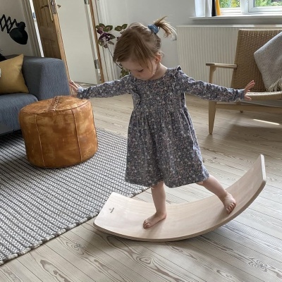 Criança de pé numa tábua baloiço de madeira clara numa sala com pufe e cadeira